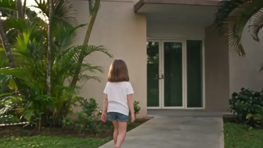Menina de costas caminha em direção à entrada de um prédio, entre plantas.