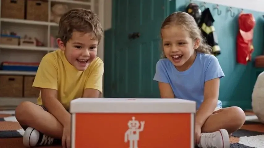 Duas crianças rindo diante de uma caixa laranja, num quarto de brinquedos.