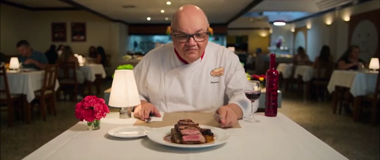 Chef de dólmã branca corta uma carne no ponto sobre a mesa de um restaurante, com o salão ao fundo.