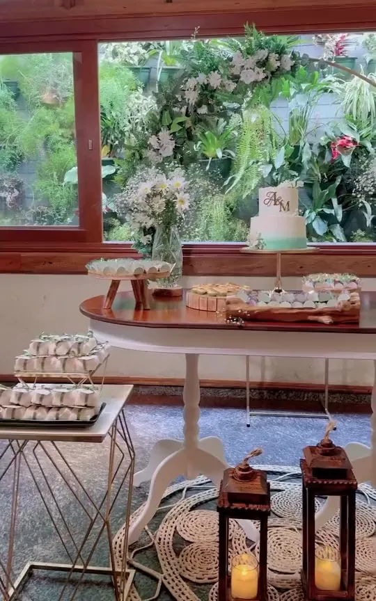 Mesa de bolo e doces montada para uma comemoração no salão da Alameda Santos, com o jardim ao fundo.