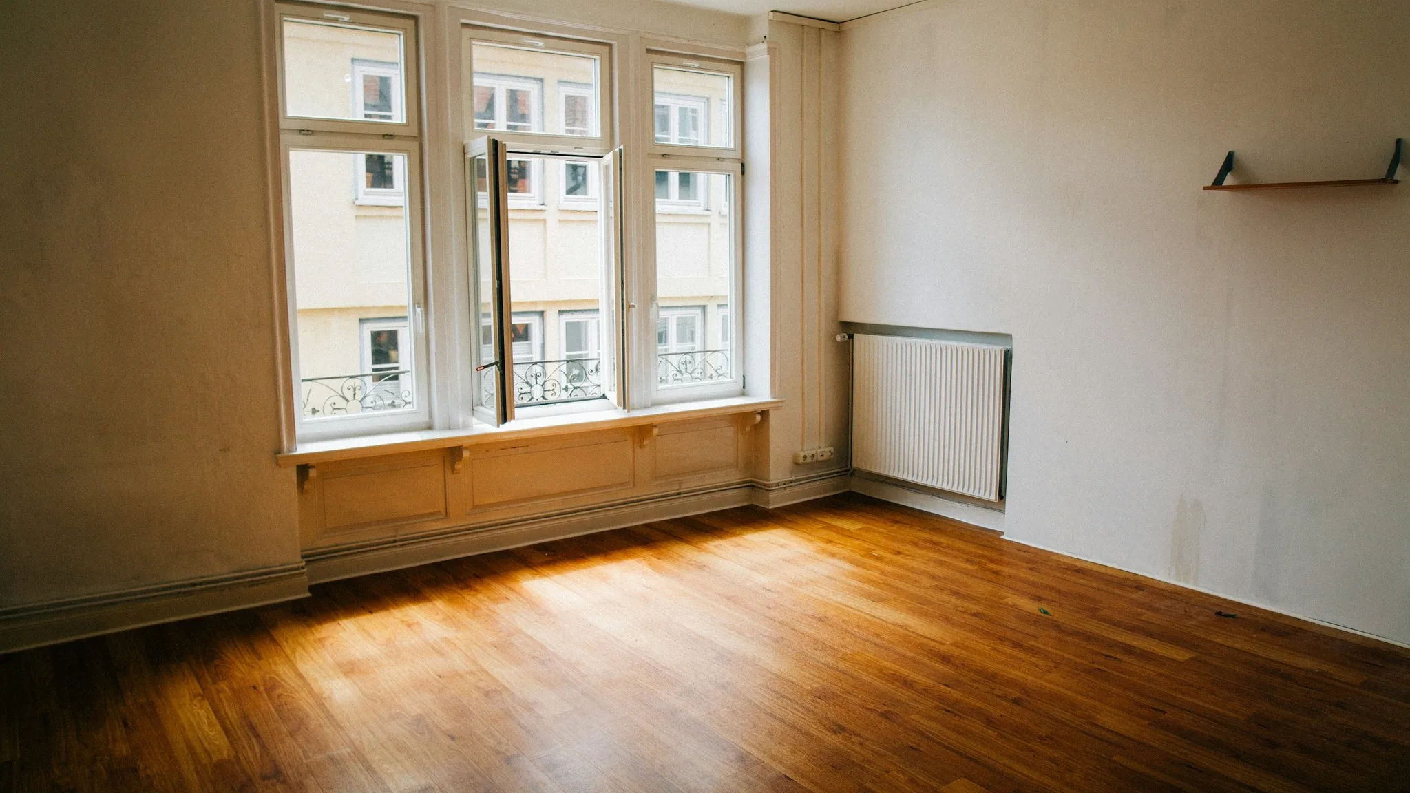 Sala vazia de verdade: piso de madeira, janelas altas e aquecedor sob a janela.