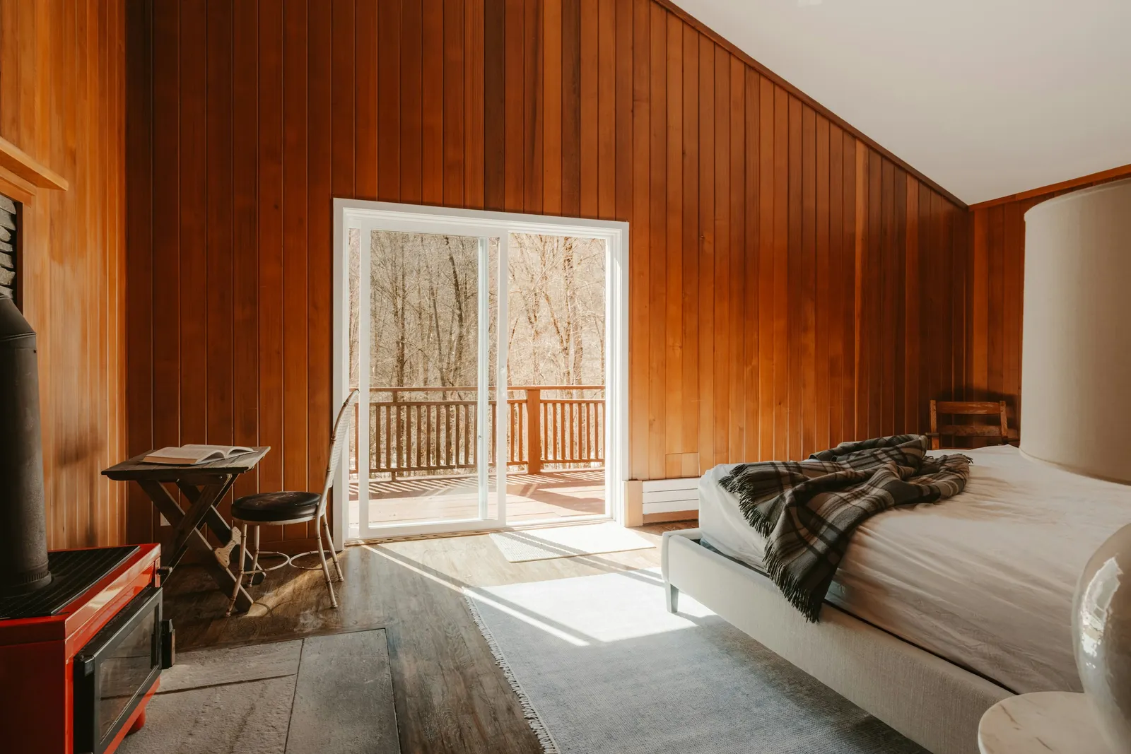 Quarto revestido de madeira, com a cama e a porta de vidro aberta para o deck.