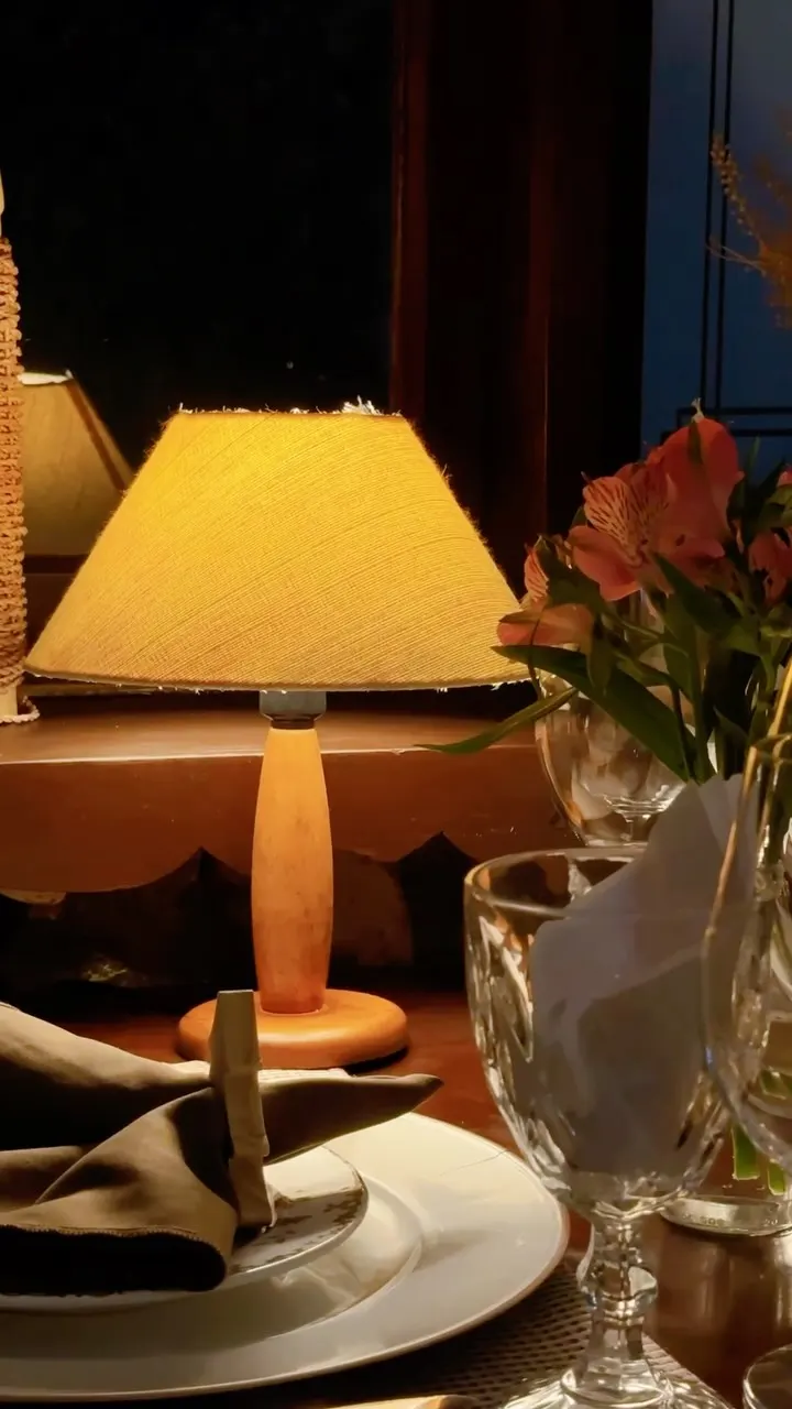 Abajur aceso sobre a mesa, com flores e taças, à noite.