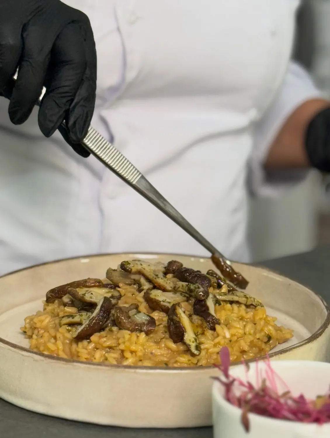 Mãos finalizando um risoto com cogumelos usando pinça.