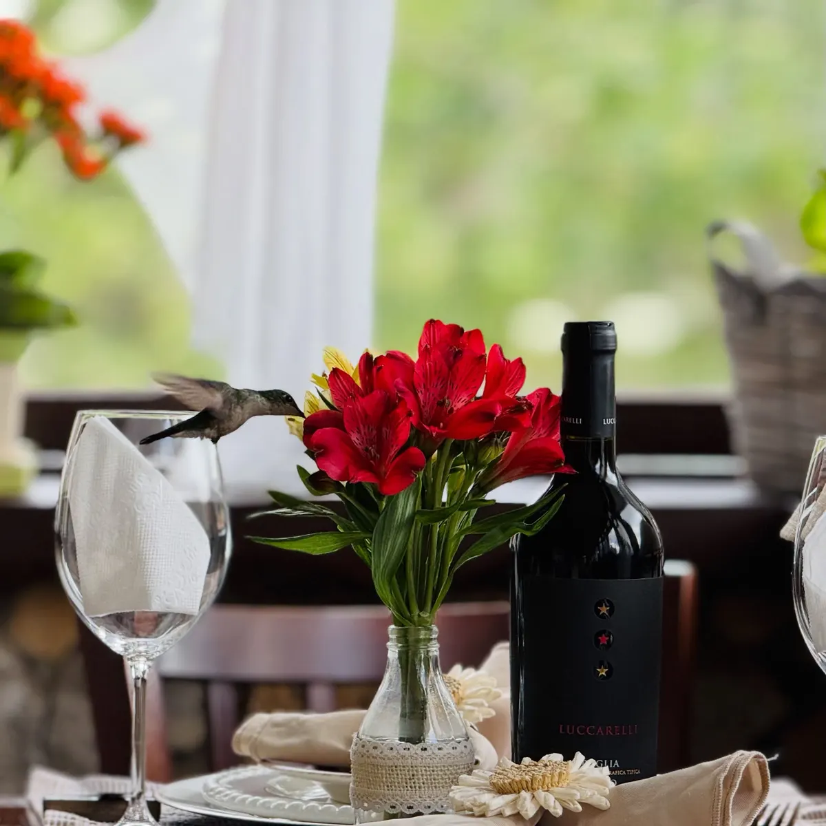 Mesa com flores vermelhas, taça e garrafa de vinho junto à janela.